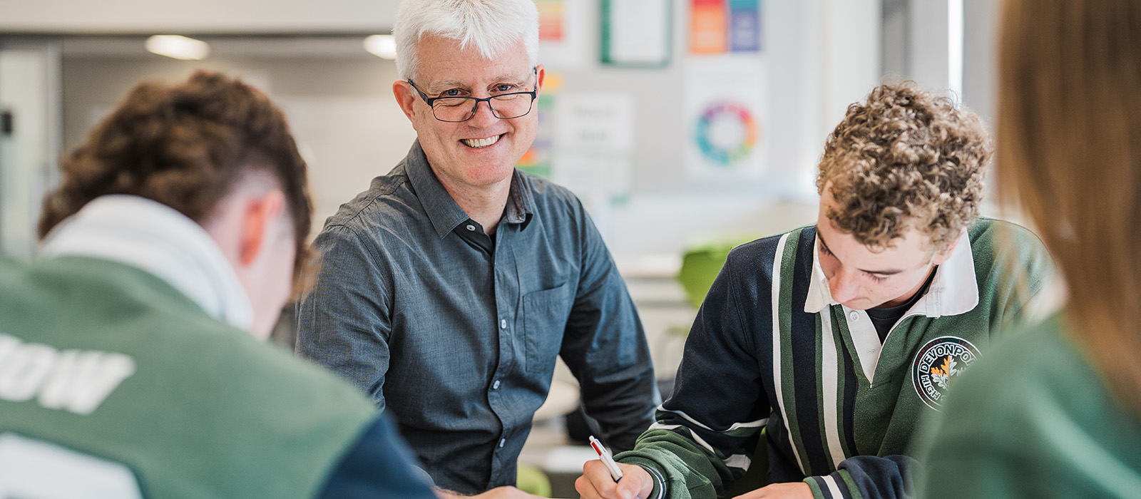Mature teacher with white hair wearing glasses smiling at the camera while students study