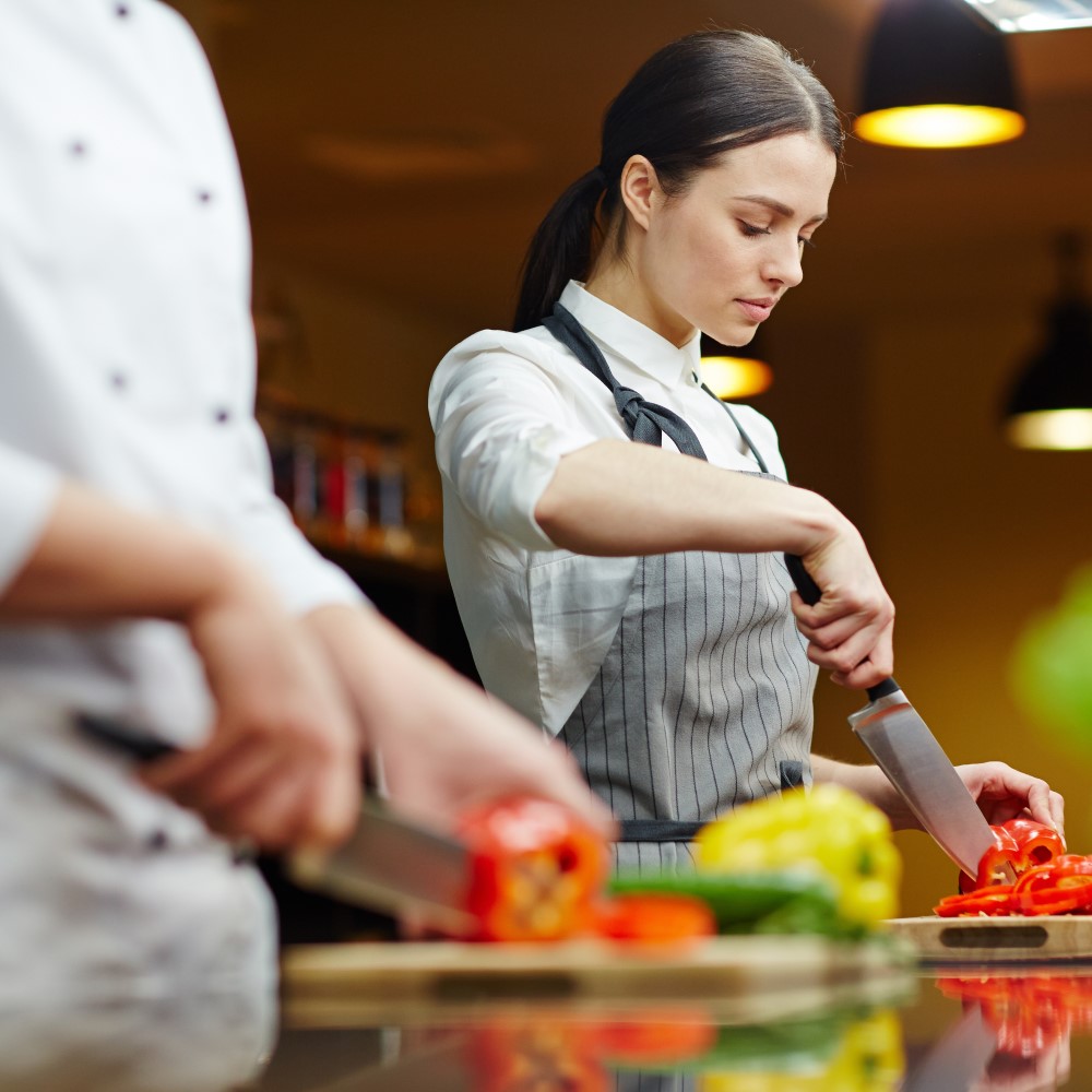 Female apprentice chef in kitchen cutting up vegetables