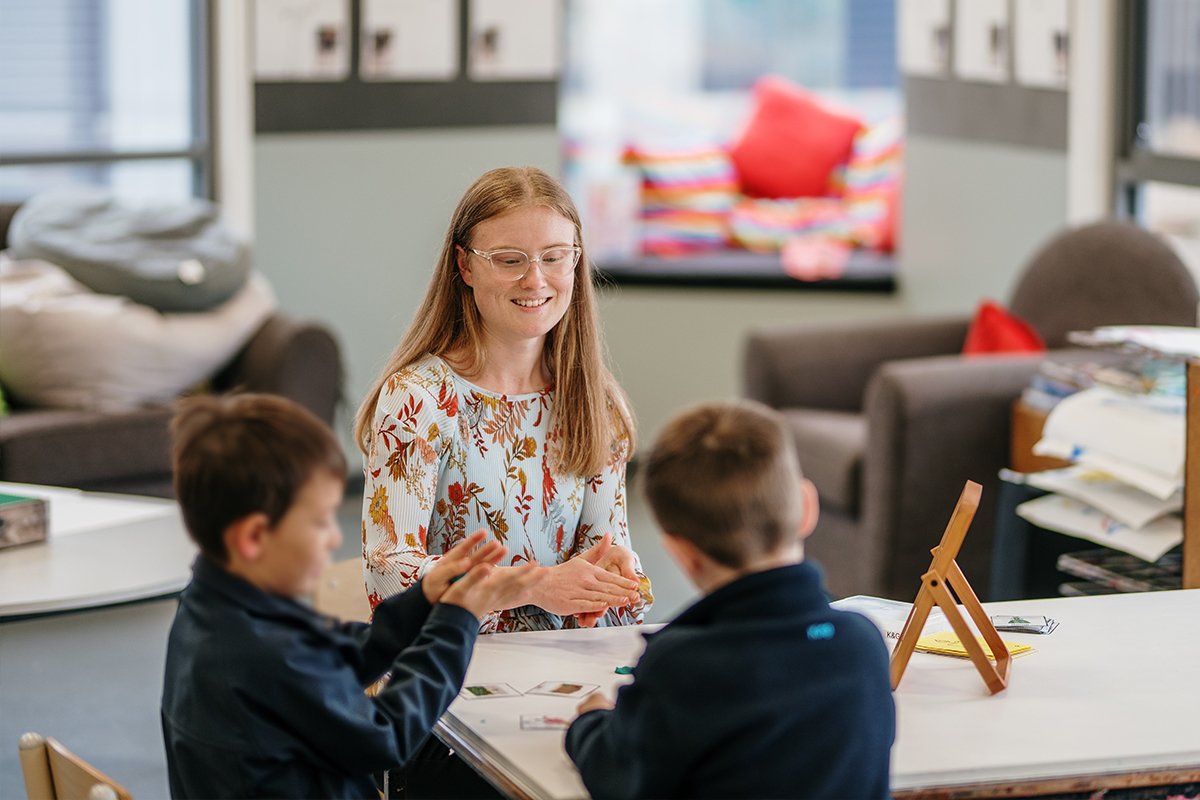 Young woman with long hair and glasses smiling and engaging with two young students in classroom