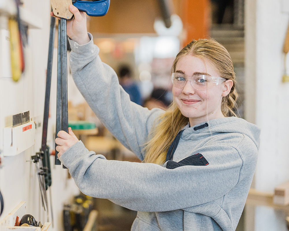 Teenage female student hanging up carpentry clamps on wall and smiling at the camera