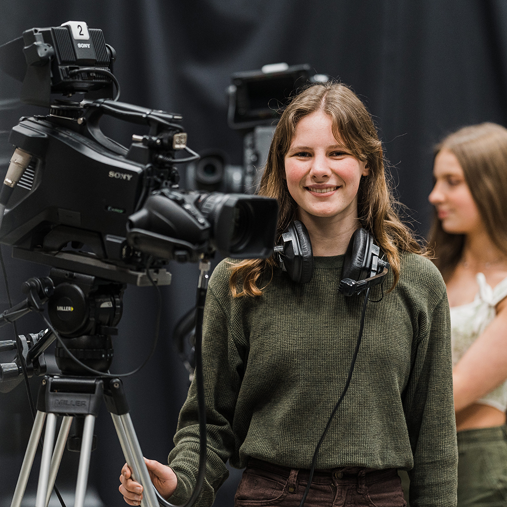 Young female student smiling and standing next to large video camera in a studio