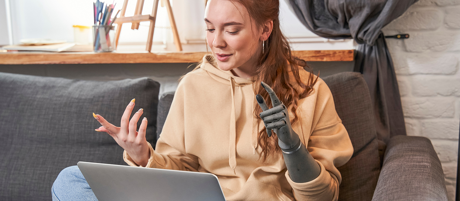 Smiling girl with prosthesis hand sitting on couch at home and talking with friend or family. Ginger girl having video call on laptop. Woman communicate with pleasure emotions, while using dating online application on computer