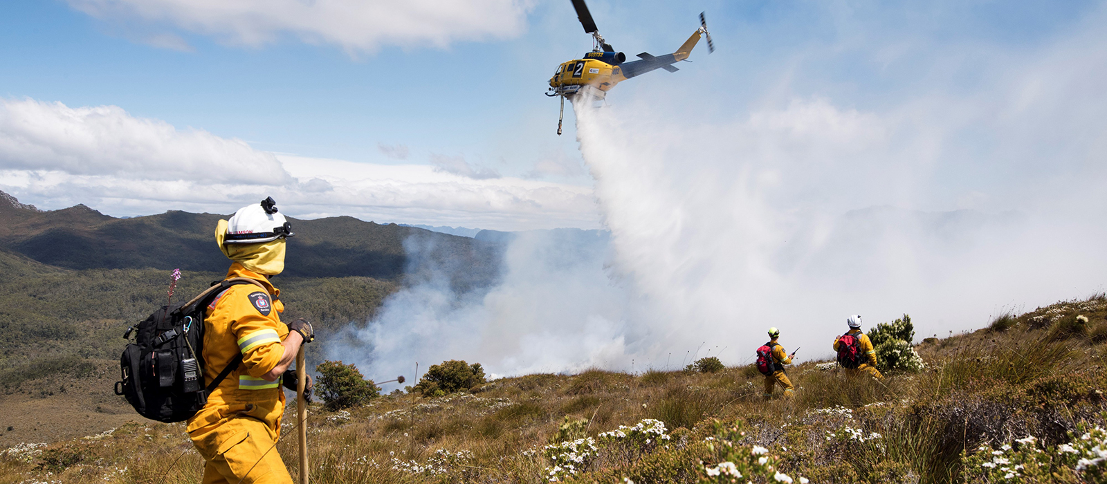 Firefighters in bushland watching a helicopter dump water on a fire