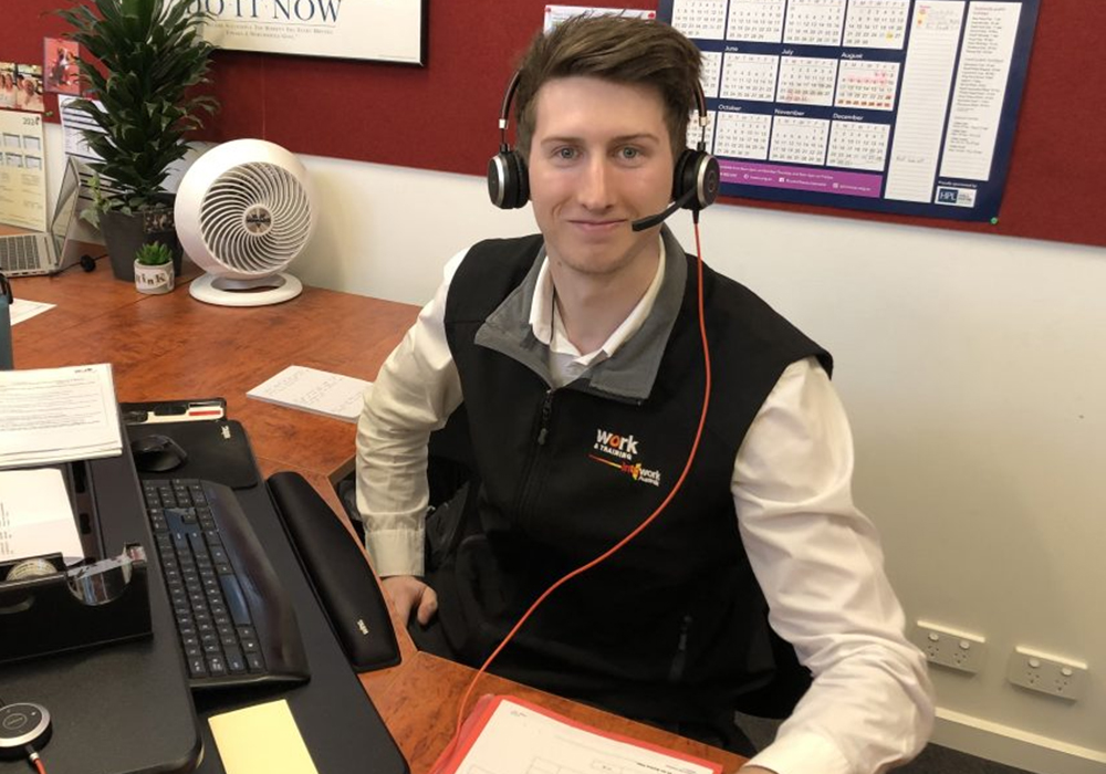 Young male trainee sitting at desk with headphones on smiling at camera