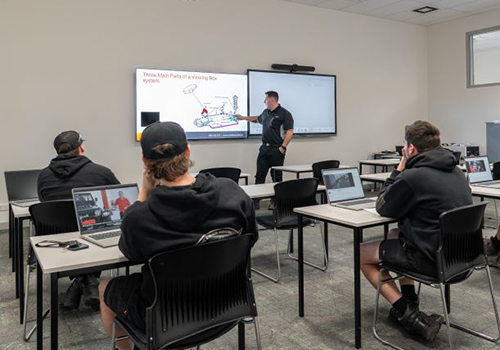 Inside of an automotive training facility building showing a number of students sitting on chairs and a teacher showing an image on a large screen